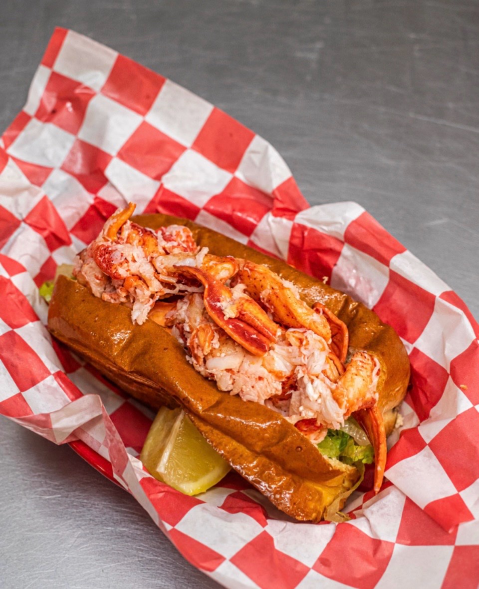 A Maine lobster roll piled with fresh claw and knuckle meat on a toasted brioche bun, served in red-checkered paper
