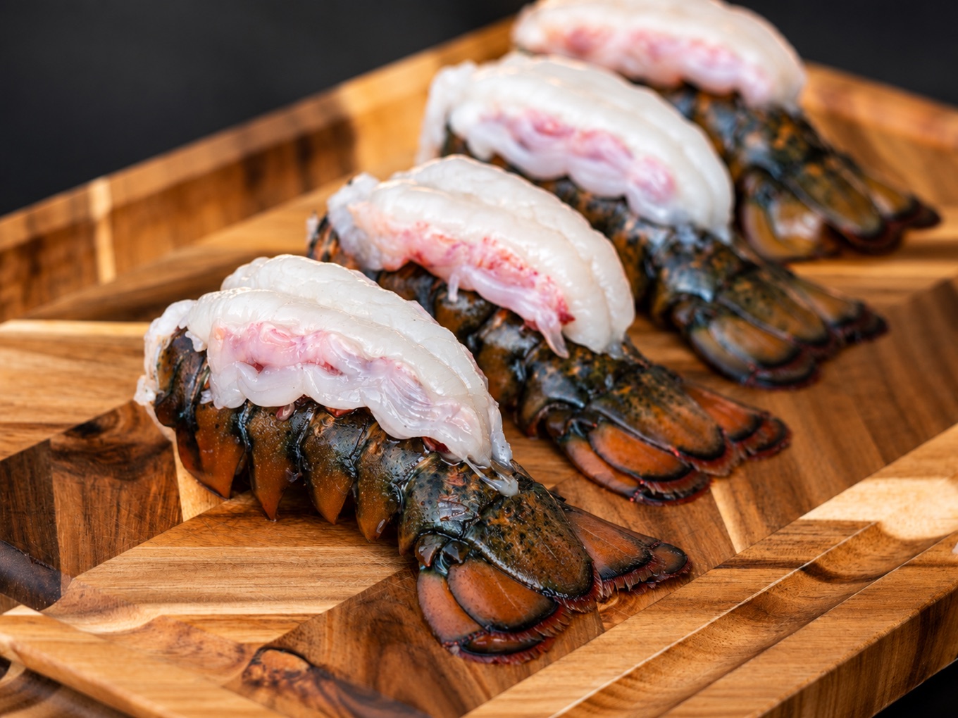 Four raw HPP shucked Maine lobster tails, the meat lifted whole from the shell, lined up on a wooden board